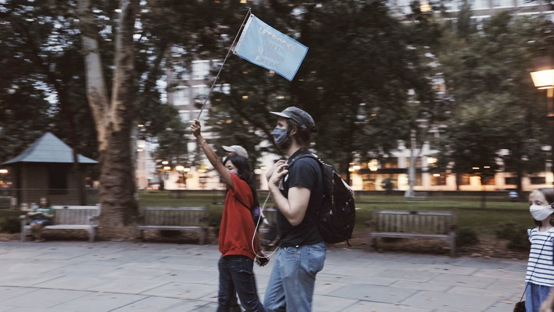 A central masked figure carrying a flag is followed by other processioners on a brick, tree-lined street