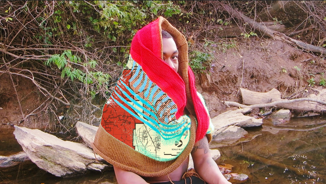 A person with short hair and dark skin makes eye contact with the camera while sitting on a riverbank wrapped in a knit textile hood with an image of a map inlaid within the fiber