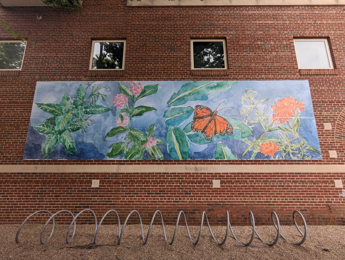 watercolor billboard depicting butterflies, flowers, and leaves on blue background against brick wall