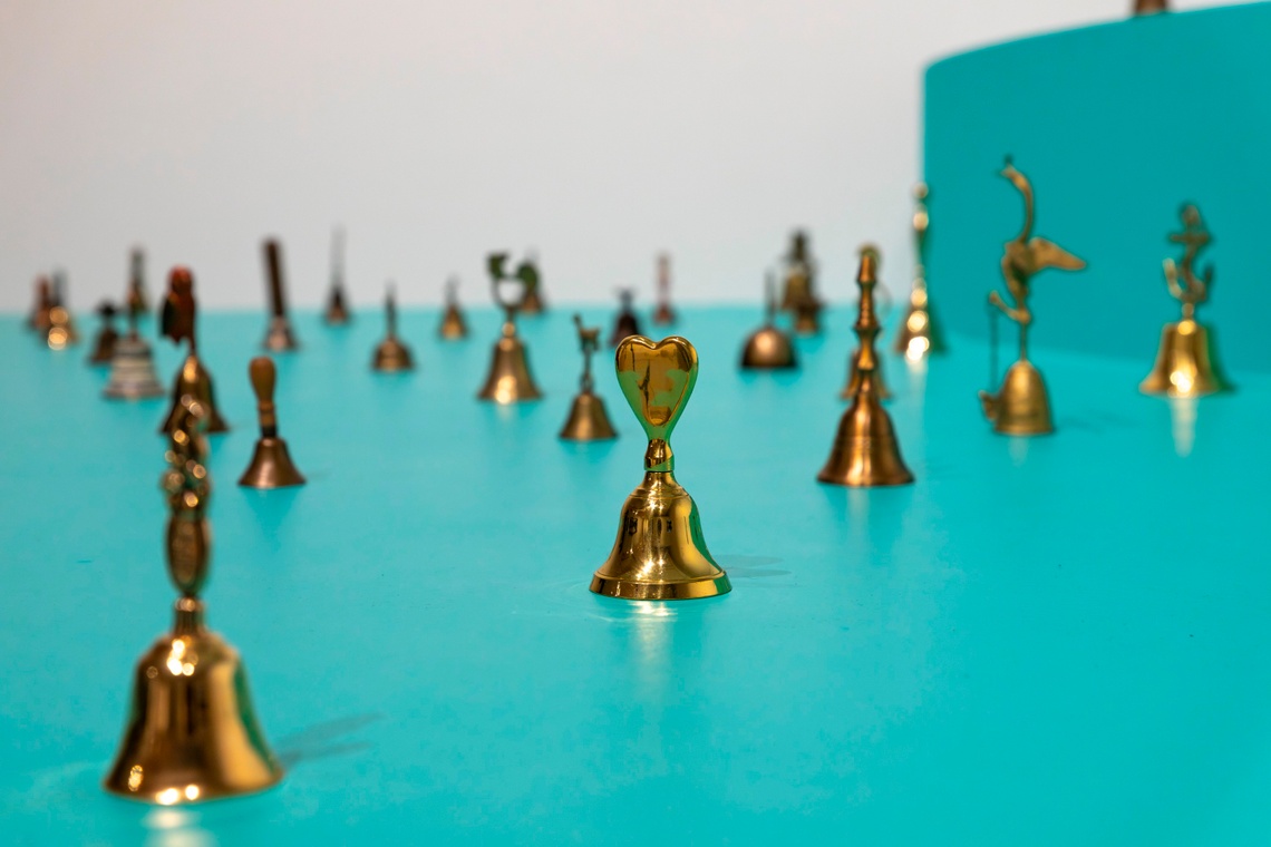 Gold bells of various shapes and sizes, with a bell topped by a heart shaped handle in the center, are arranged evenly on a turquoise platform