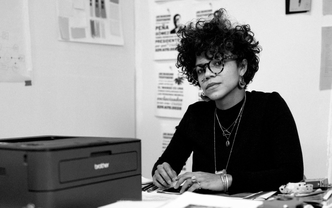 A black and white photograph of Cruz at a desk by an inkjet printer and a stack of papers