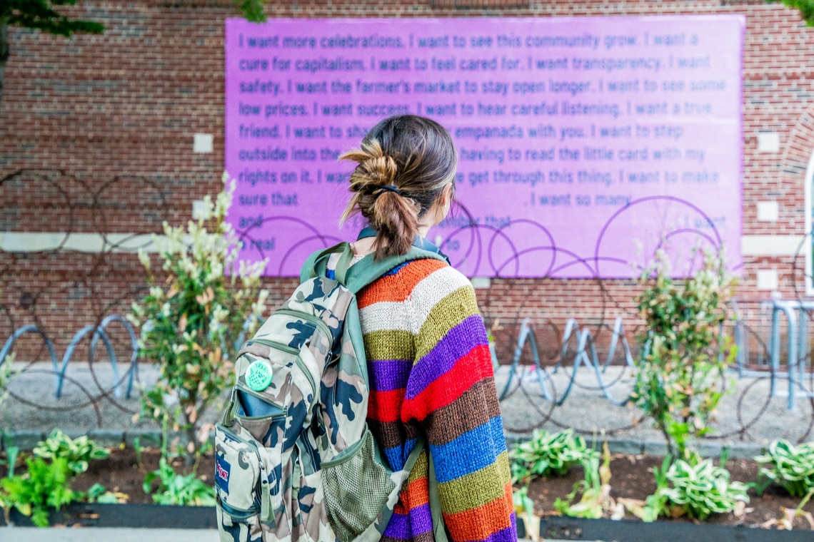 A woman in a striped sweater looks at a pink billboard with green text that is out of focus.