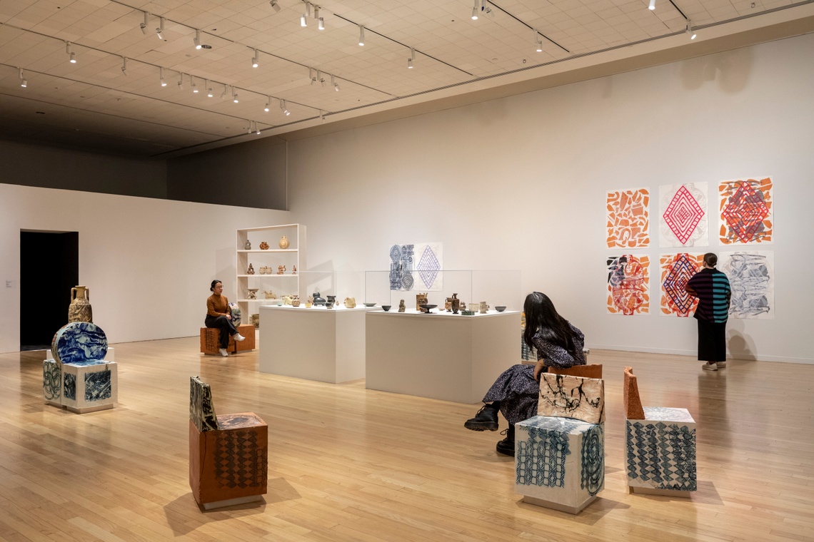 three people engage with artwork installations looking at hanging prints small sculptures and ceramic seats
