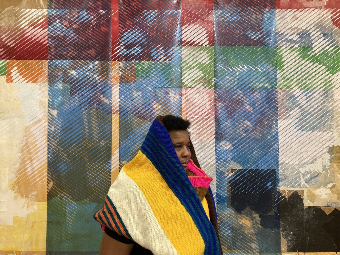 A woman sits in front of a screenprint photo collage wearing a multicolored knit hood