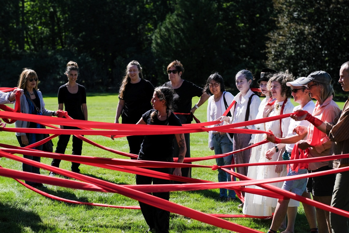 A group of people connected by a pink ribbon in a grass field
