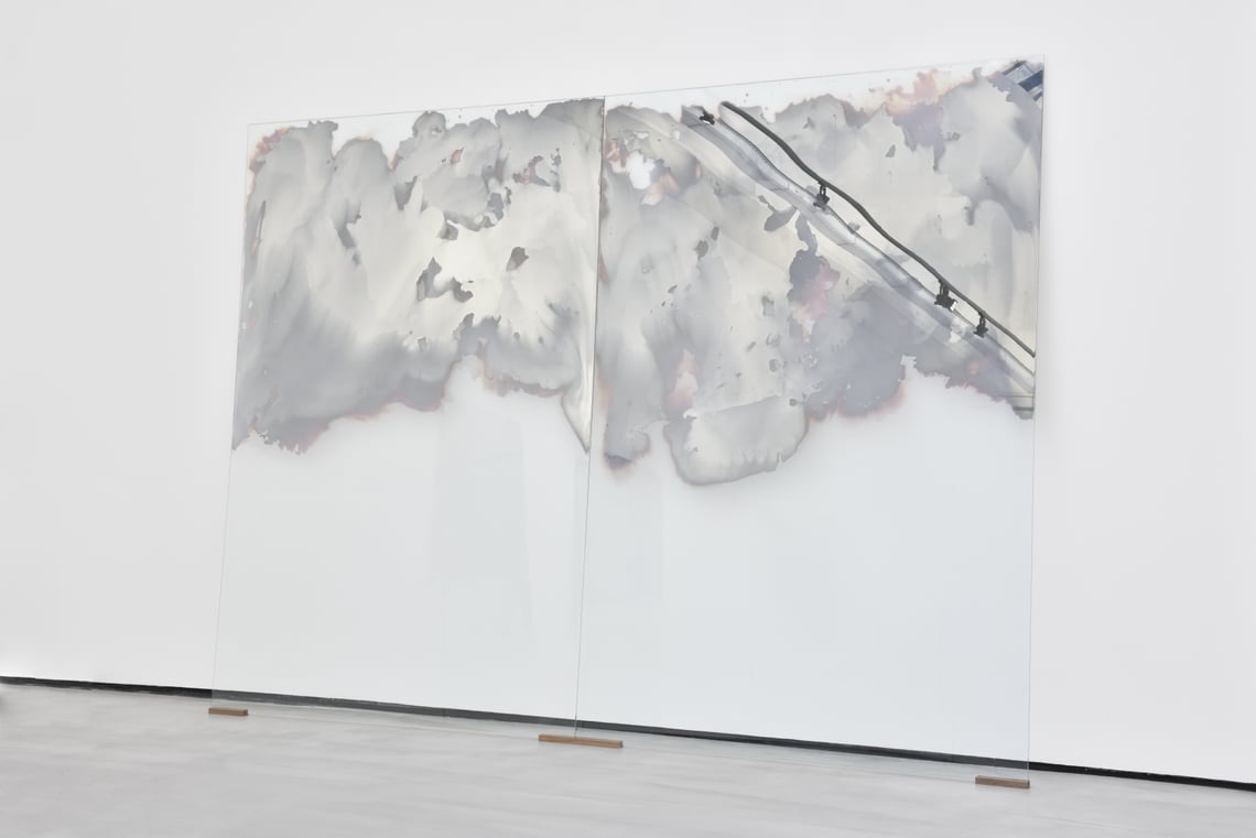 Grey cloudy forms billow from the top of two glass panels leaned against a wall