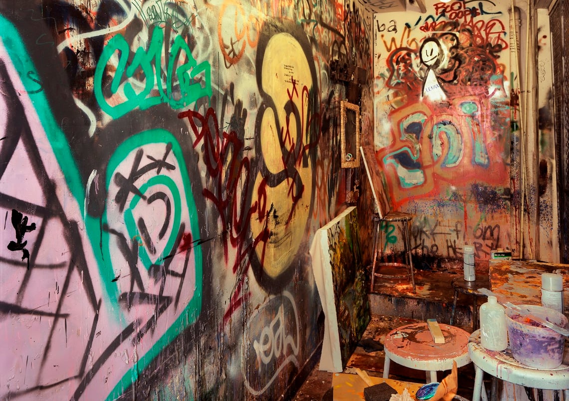 A narrow room filled with multi-colored graffiti with a canvas leaning on the wall next to a short artist stool