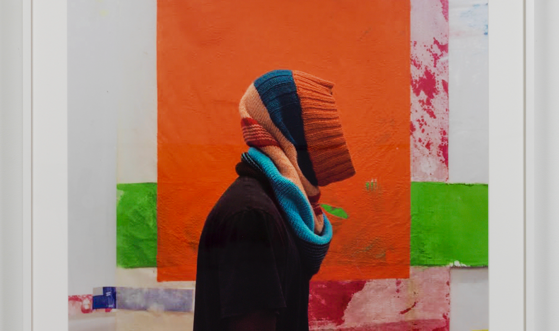 A photograph of a striped knit hood made green, black, and red yarn in different shades mounted in a white frame