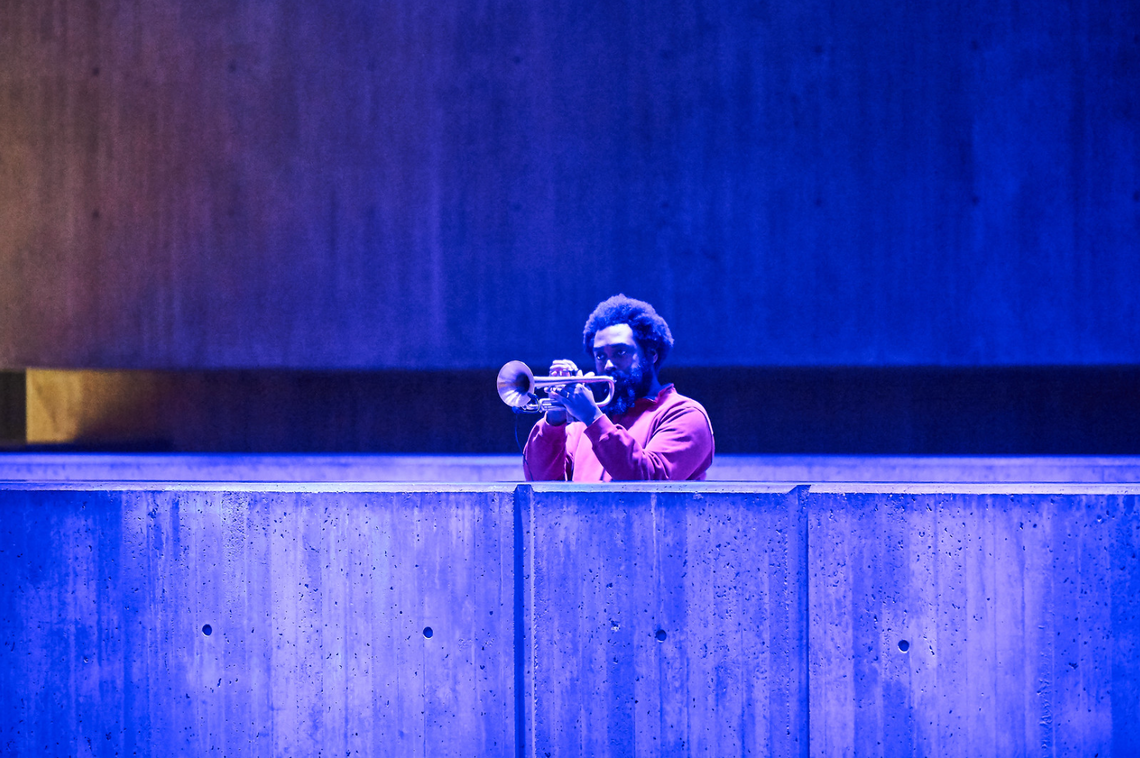 Forbes Graham plays a trombone lit by pale blue and purple light behind a wall on a stage