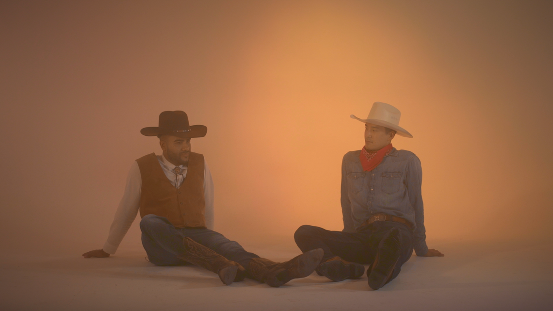 two male figures in cowboy hats and outfits sit with legs outstretched against a hazy yellow tan studio backdrop