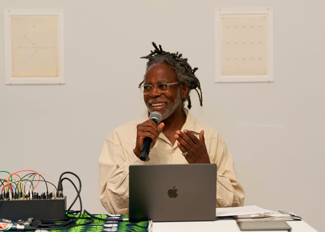 Arnold J Kemp, a man with locs and glasses, smiles while speaking into a microphone. He is seated at a table with a laptop and audio equipment, gesturing with one hand. Two framed artworks hang on the wall behind him.