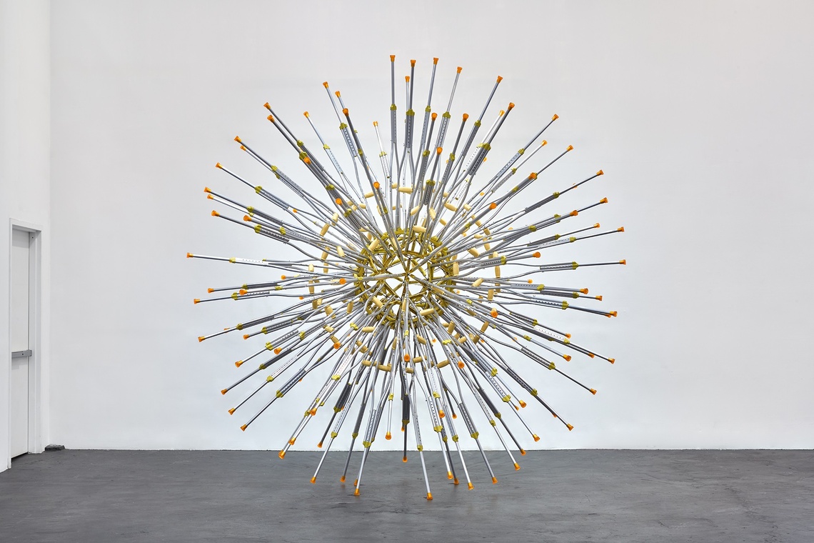 Yellow-rubber topped crutches are suspended in a starburst arrangement in a white room with dark gray concrete floor