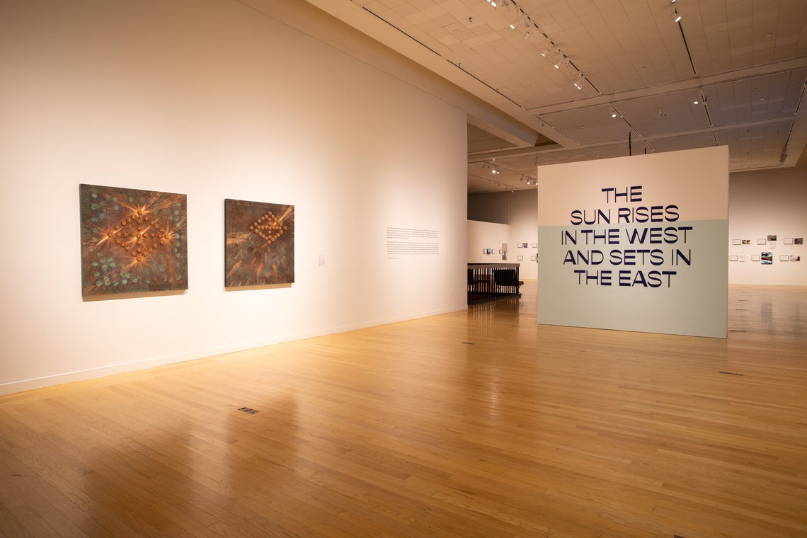 Installation view of The Sun Rises exhibition with large white walls and two copper artworks on the left wall
