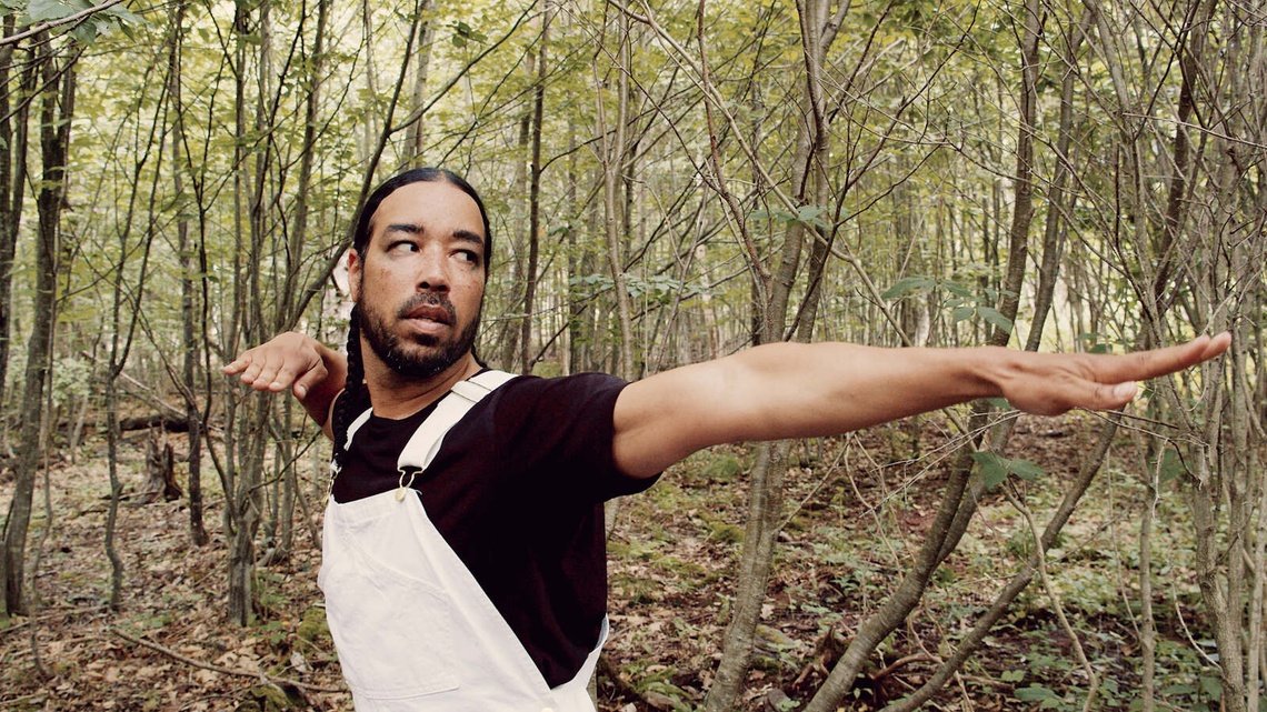 A person in white overalls in a green wooded area dramatically reaches out their left arm