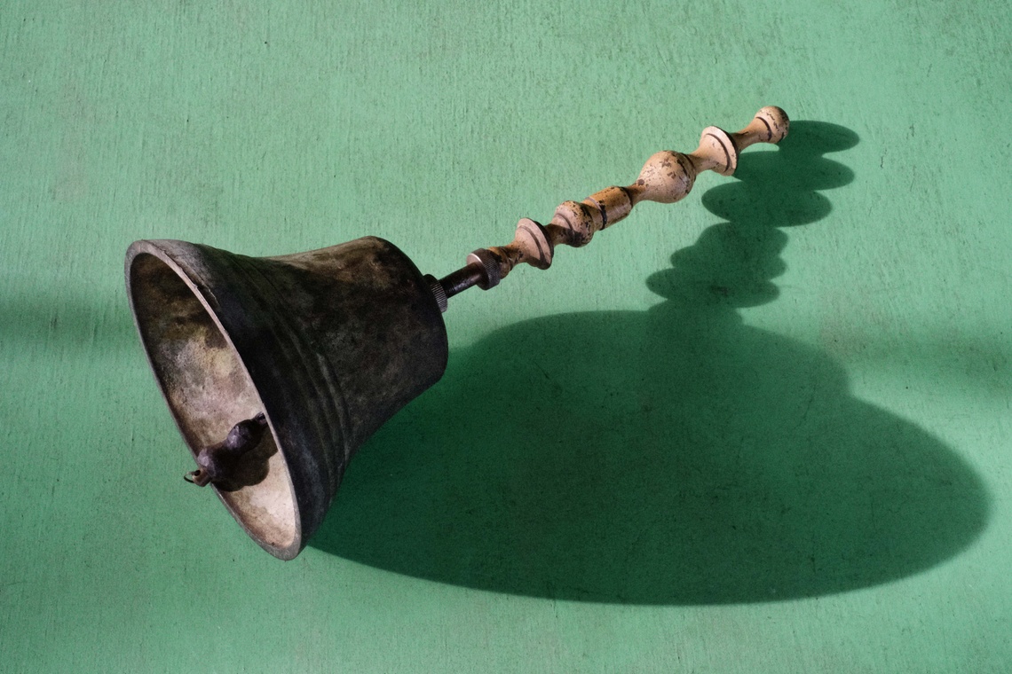 An ornate antique bell with a carved wooden handle casts a shadow on a green surface
