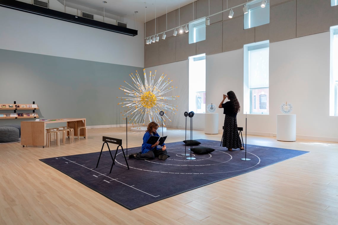 Two women engage with an installation on a black carpet in a room filled with natural light—one is seated, reading a guide book, and the other is holding a phone to her ear listening to an audio guide