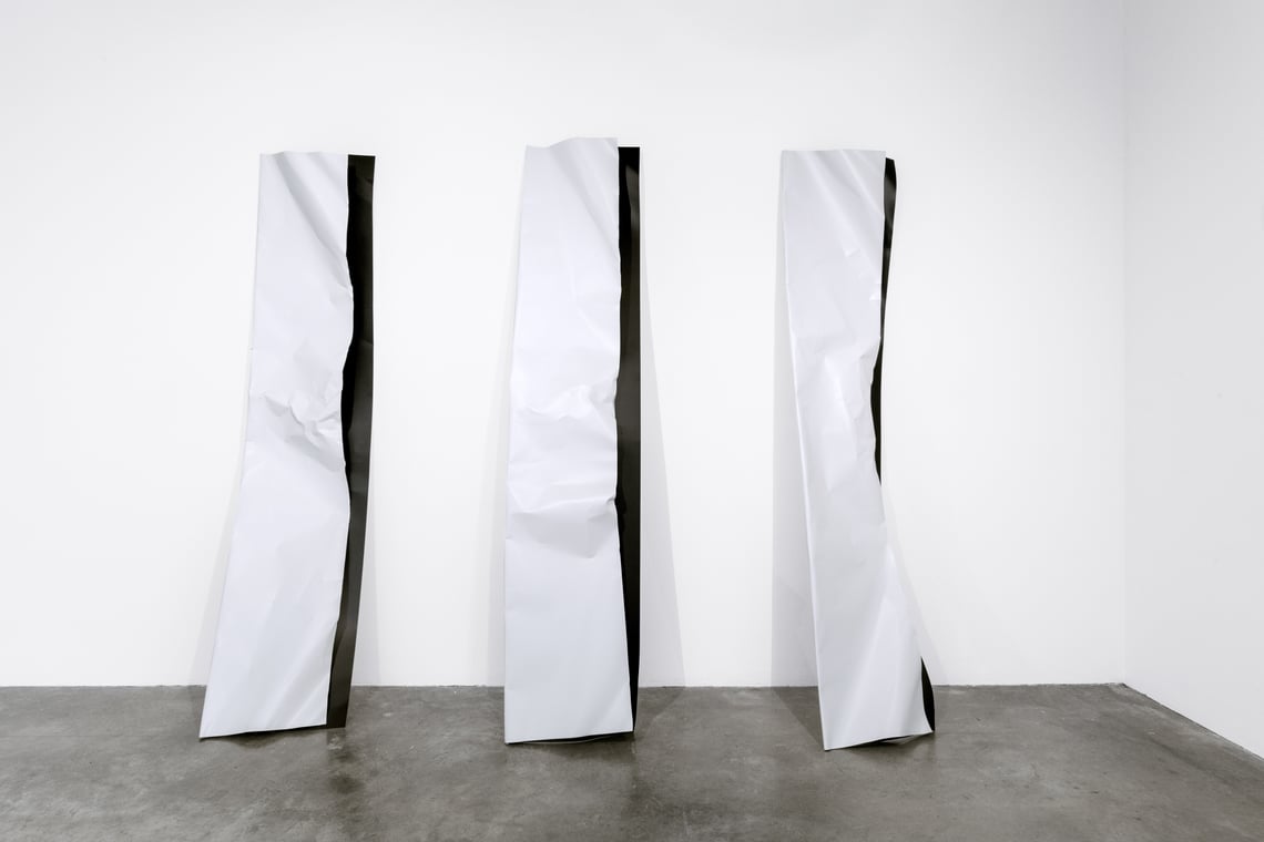 Three crumpled white metal planks lean irregularly against a white wall on a concrete floor