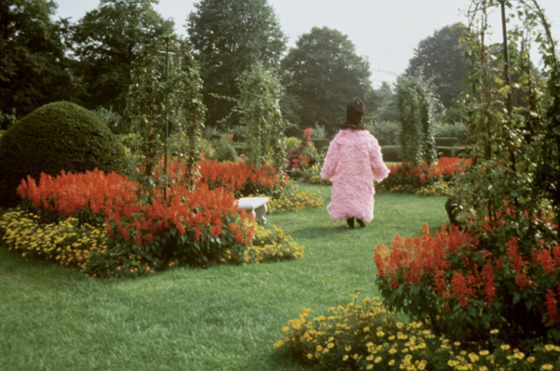 A figure in a pink organza ruffled coat with a black shroud walks through a green garden surrounded by flowers