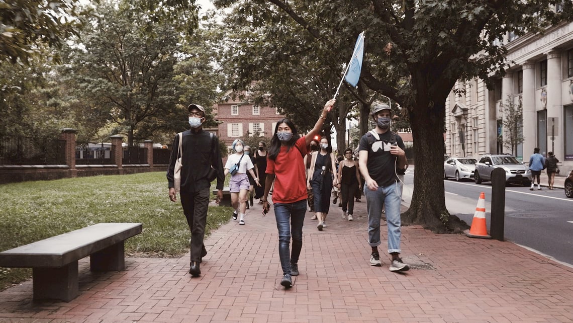 A central masked figure carrying a flag is followed by other processioners on a brick, tree-lined street