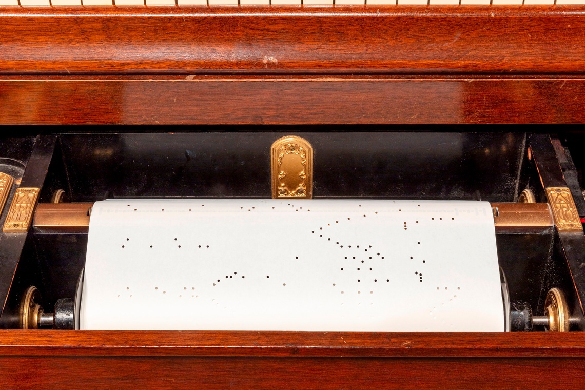 A player piano scroll, white with irregular hole punches, is rolled into a cherry wood piano.