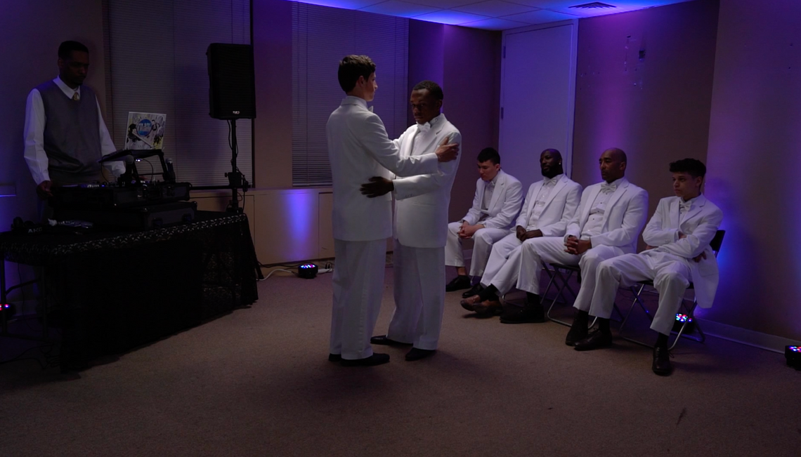 men in white suits sit watching two men dance in a dimly purple lit room
