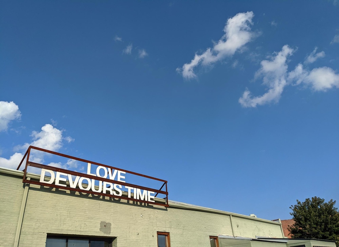 A large iron frame is attached to the roof of a low concrete building with white text reading "LOVE DEVOURS TIME" under a blue sky with a few white clouds.