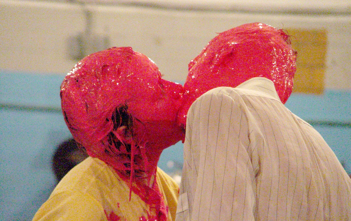 Two people with their heads and faces covered in pink paint kiss and embrace each other