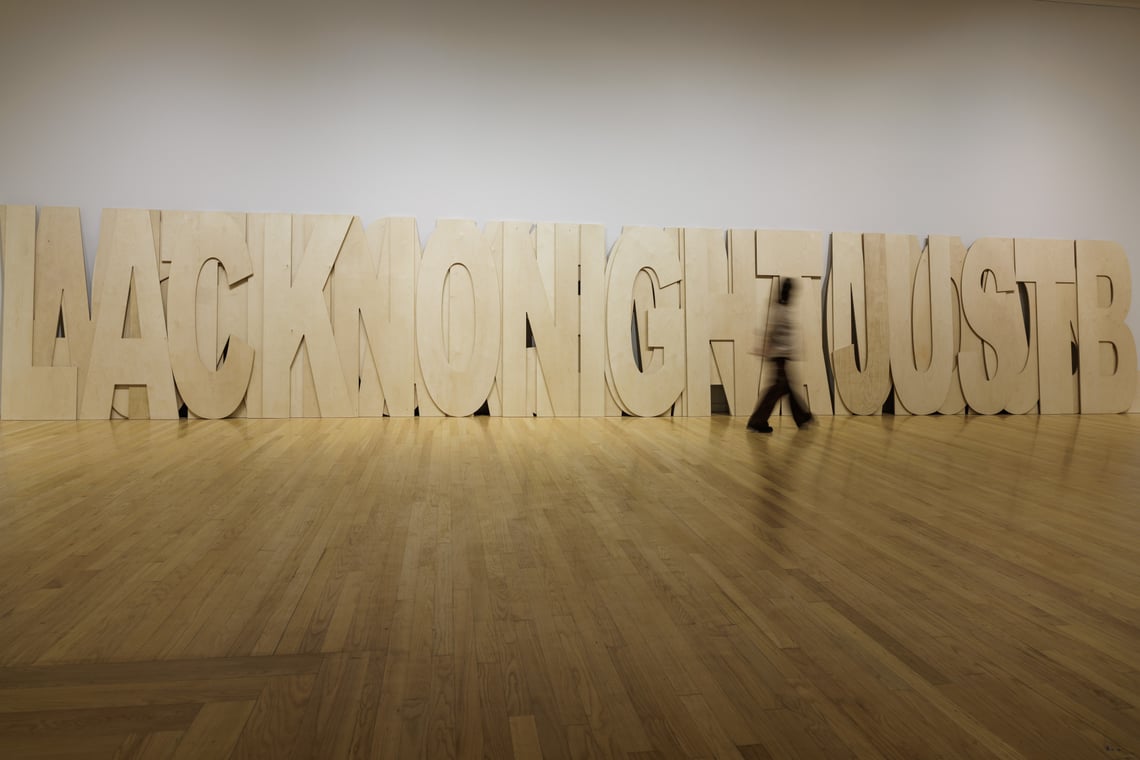 A blurred figure walks by a collection of large light wood plywood ietters leaning against a white wall in a gallery space.