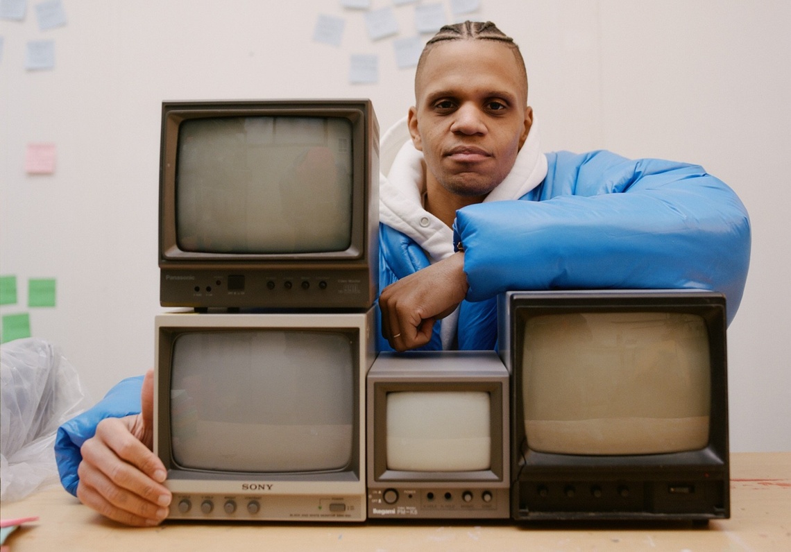 A person with tight braids wearing a bright blue puffer jacket rests their arm on a stack of old televisions while making eye contact with the camera.