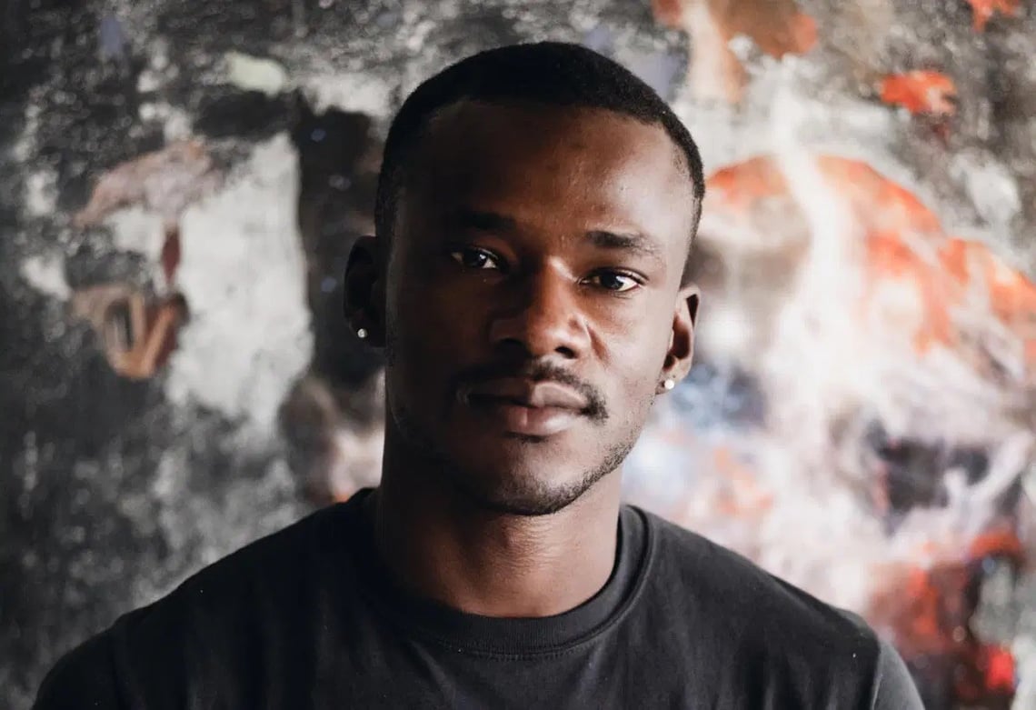 A dark-skinned man with closely shaved hair and moustache looks into the camera in front of an abstract warm-toned painting