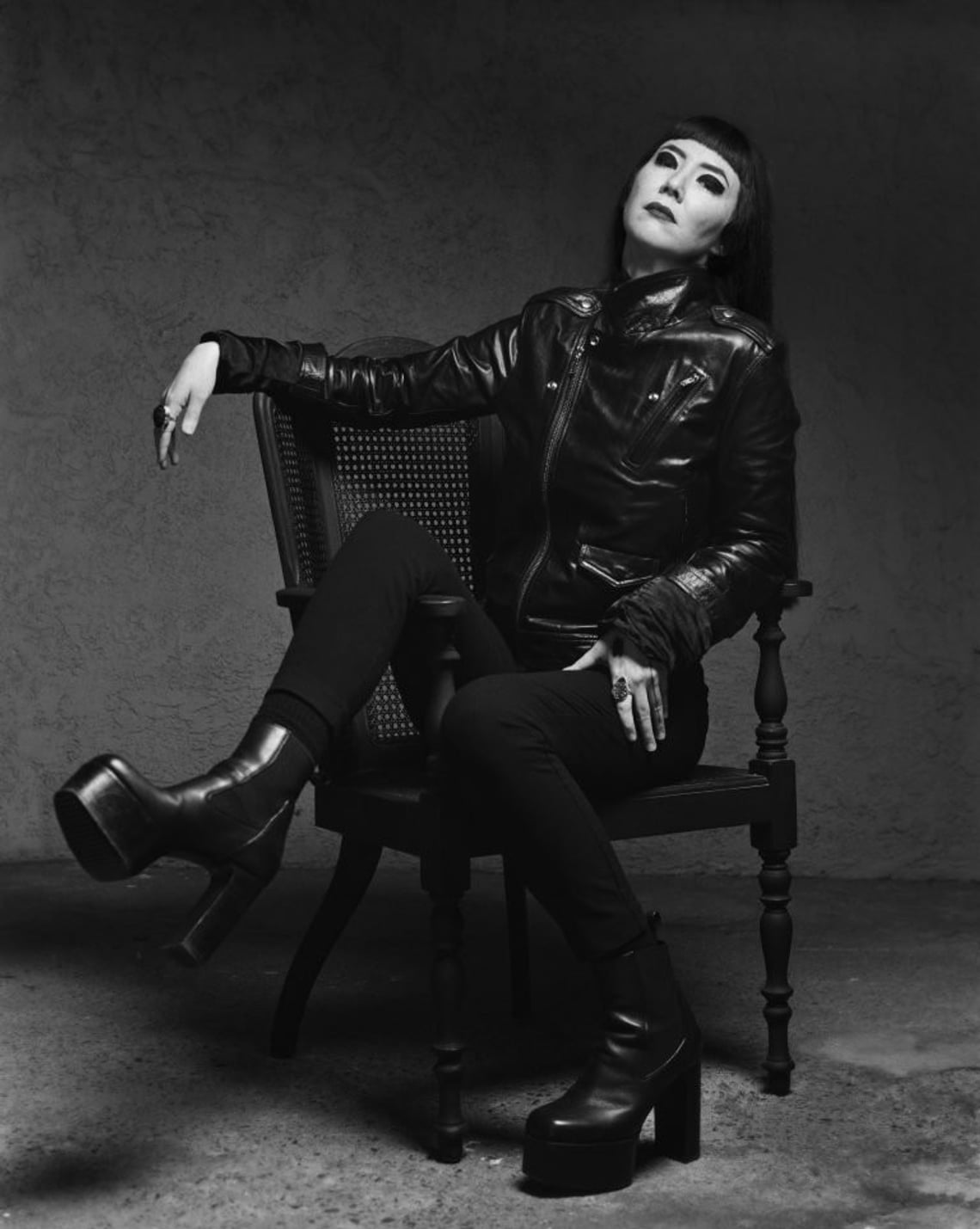 A black and white photograph of a person with long, dark hair with blunt bangs and heavy black eyeliner reclining in a cane chair while wearing a leather jacket and leather platform boots
