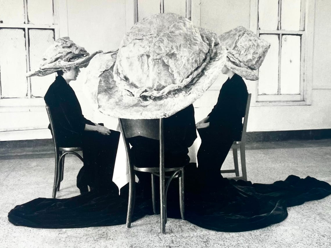 Black and white image of four cloaked figures wearing large hats sitting around a table in a windowed room
