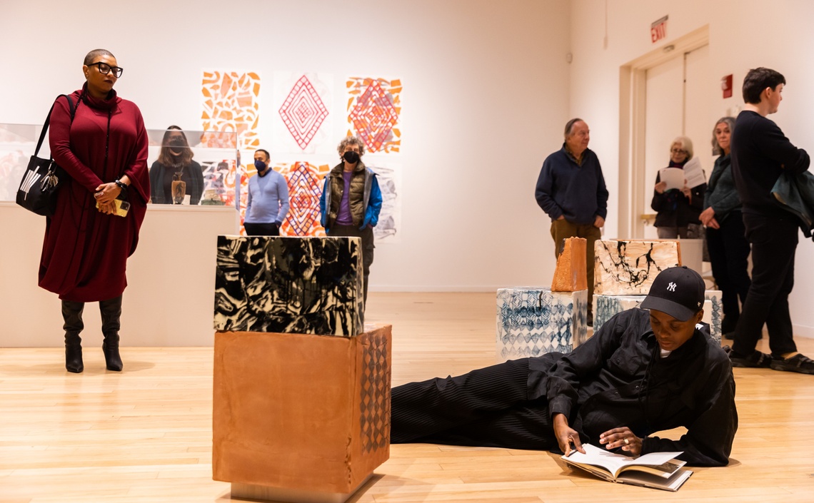 NIC kay lays on the gallery floor reading from a book as visitors watch them