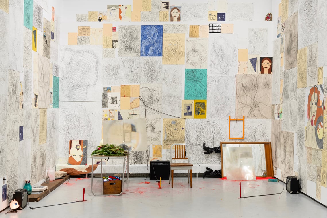 A small white room with light grey floor is covered with line drawings of a feminine face and figure on white, ivory, blue, yellow, and green paper; there are a number of objects on the floor, including a small table covered in stemmed roses laid on their side, a small chair, a microphone and two speakers, and a milk crate filled with small LED flashlights