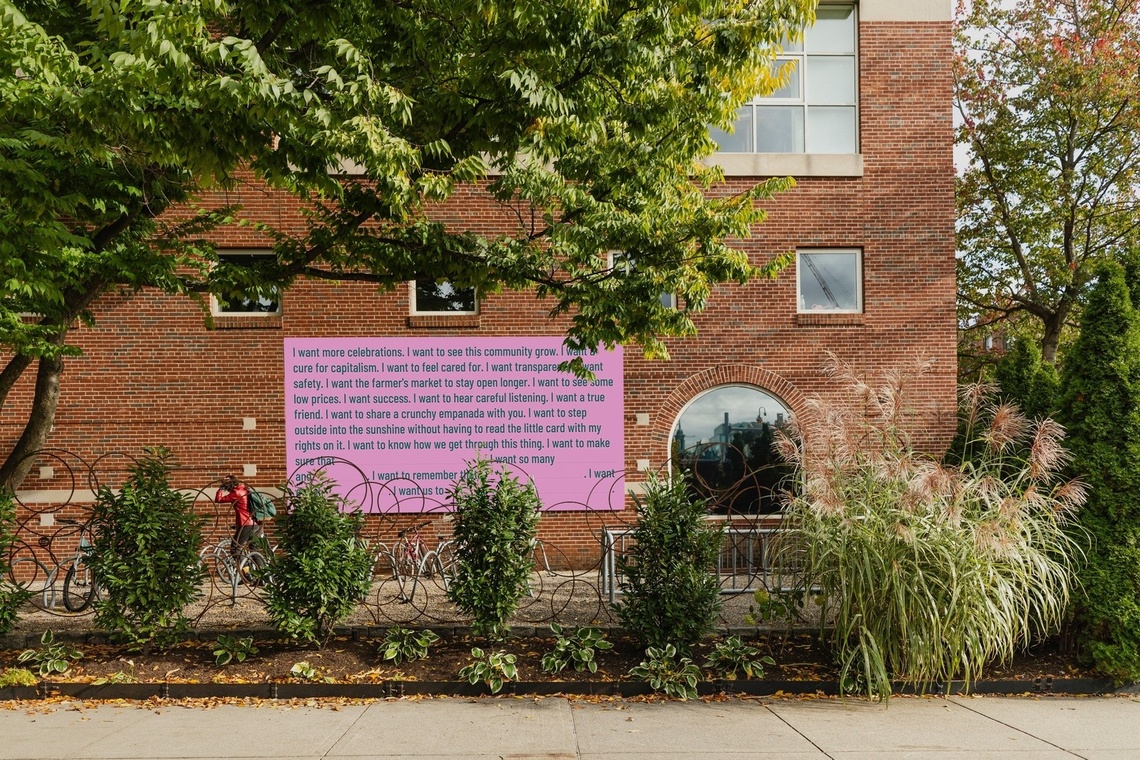 A pink billboard with green text on a brick wall surrounded by greenery reading "I want more celebrations" 