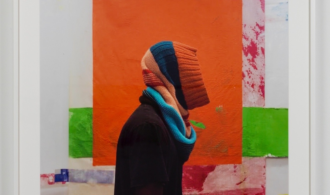 A person, whose face is made invisible by an orange, blue, and teal knit cowl, is photography from the waist up in front of a colorblocked painting.