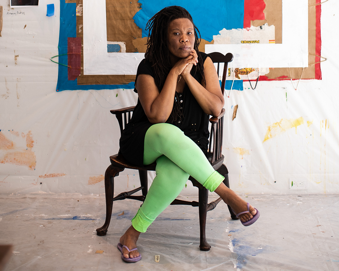 Photograph of Tomashi Jackson with long, dark locs sitting on an antique chair in front of a large brown, blue, orange, and white textile wall hanging