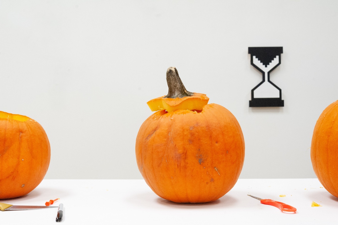 A photograph of three pumpkins in the process of being carved on a white table in front of a white wall with a hanging graphic of an hourglass