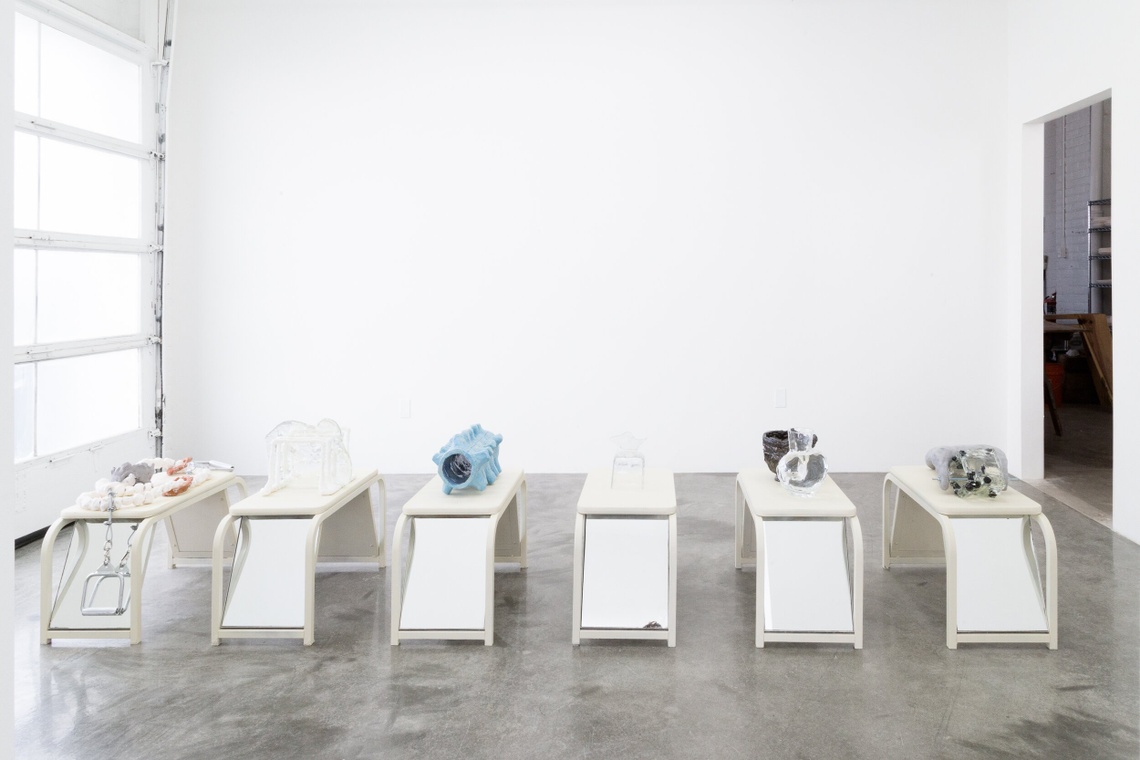 Ceramic vessels sit on top of white mirrored shoe benches in a white room with concrete floor