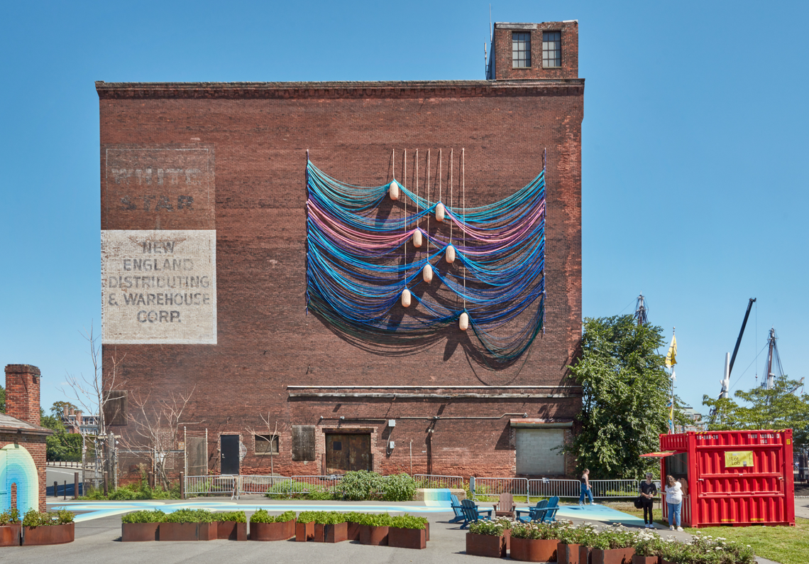 A string swag sculpture attached by buoys hangs from a brick warehouse builidng.