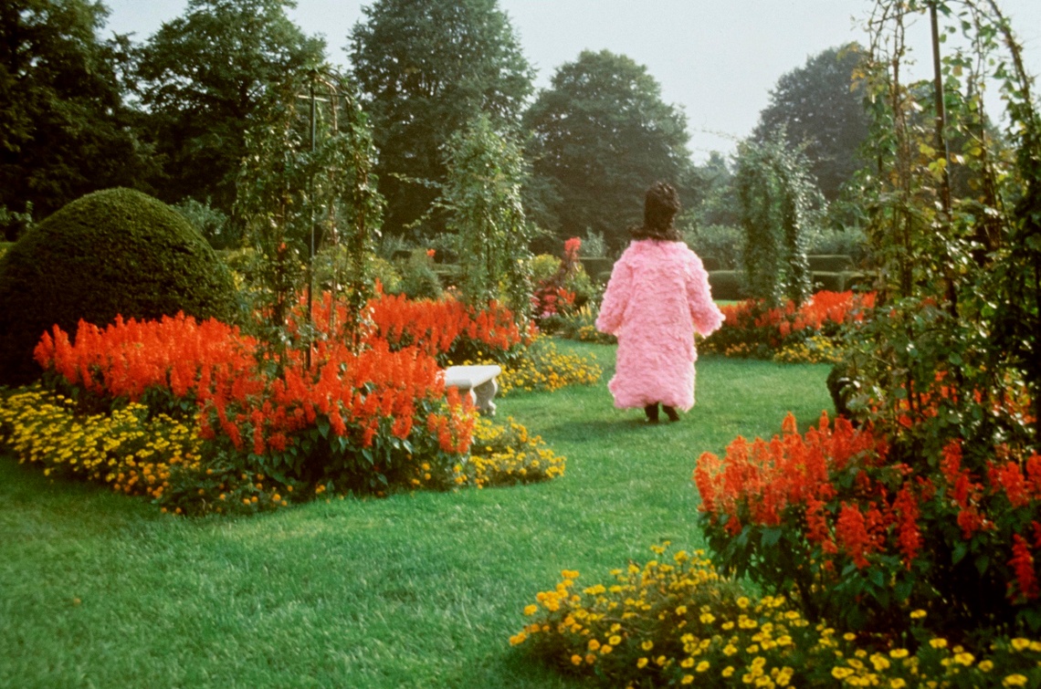 A figure in a pink organza ruffled coat with a black shroud walks through a green garden surrounded by flowers