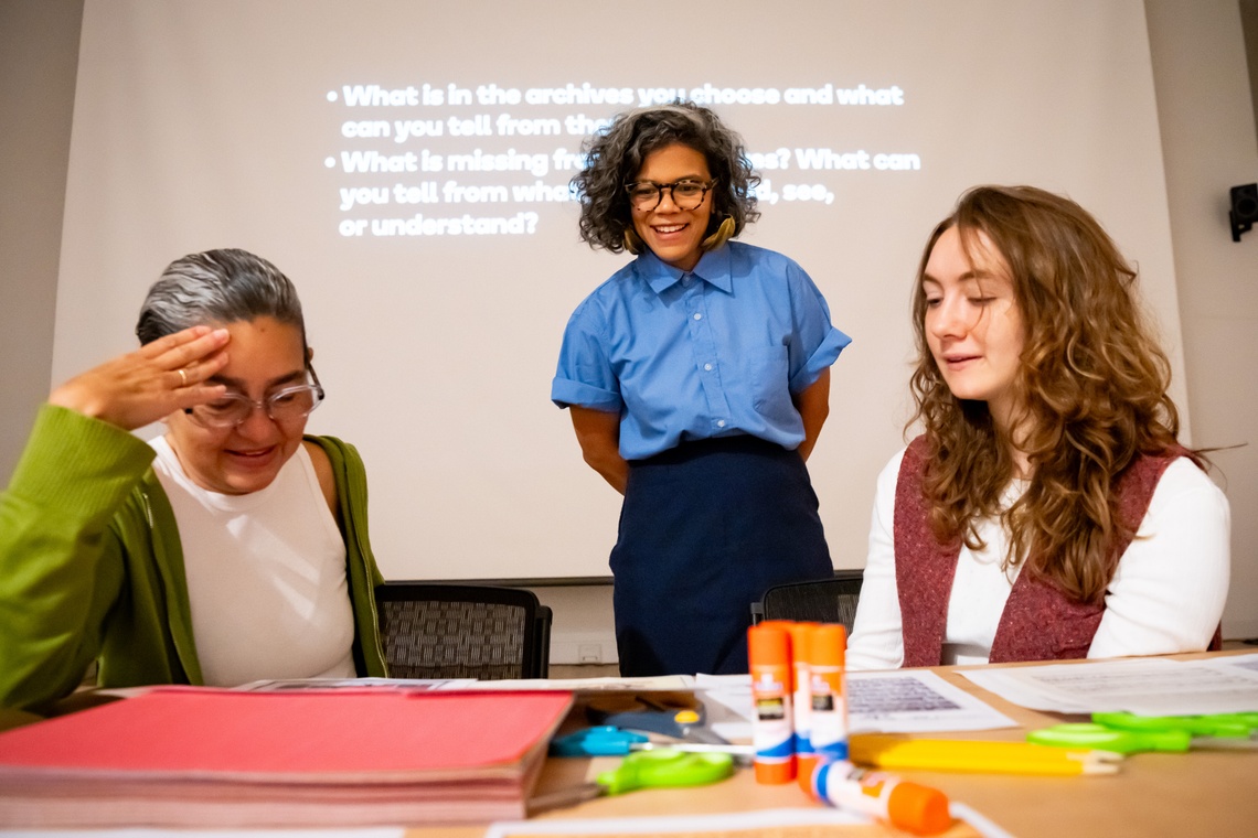 Lizania Cruz in a blue button down stands smiling behind two workshop participants at a table with paper and glue sticks.