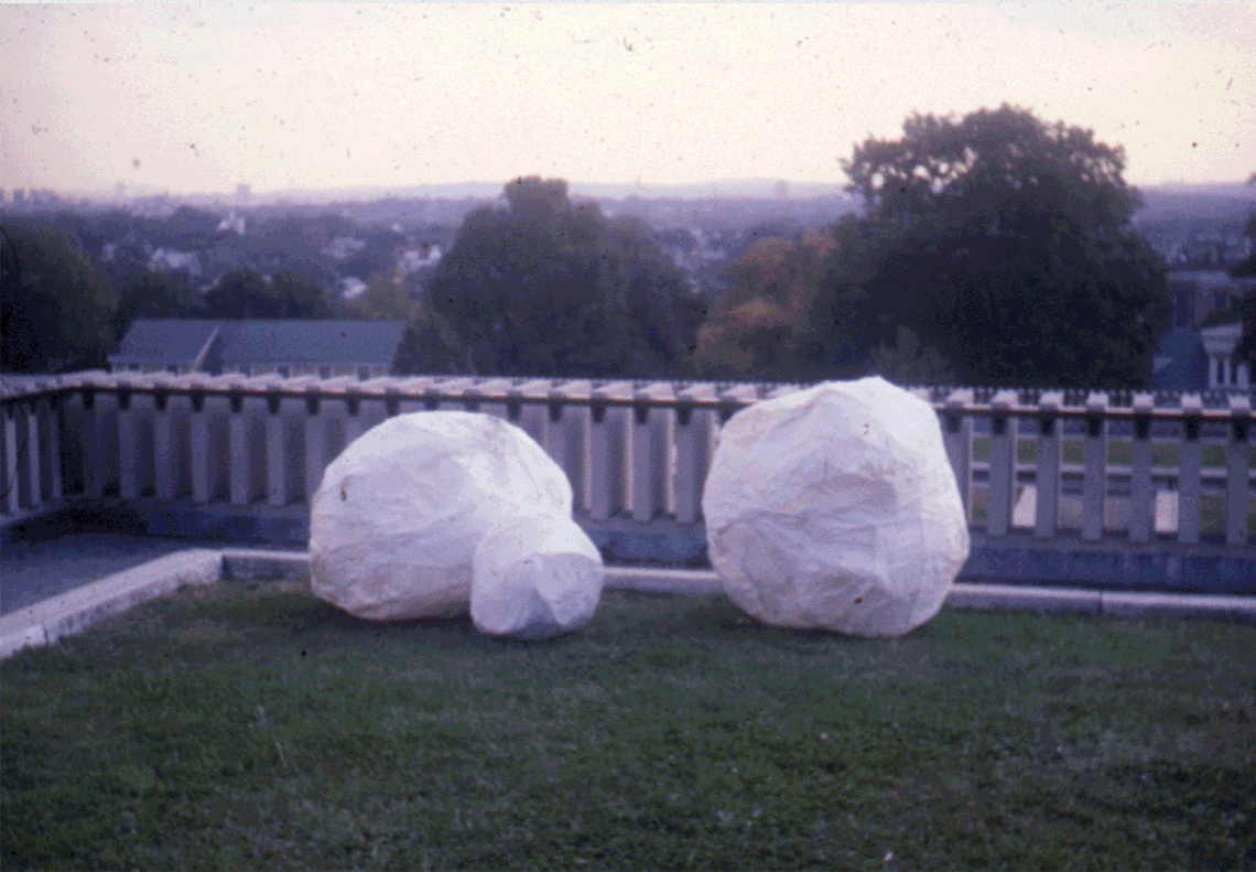 A .GIF alternating between images of three cloth boulders on a green lawn, a red fabric flag with a yellow interior, two large orange robes with photo prints hung on a wall, and shoes in pink socks with black loafers kicking matching pink potatoes